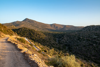 View from the road from Marathonas beach to Pachia Rachi westwards into the valley of Eleonas, an uninhabited plateau on the island of Aegina. The valley here is covered with rocks and boulders and is home to an ancient olive grove. The 532 metre high mountain Hellanion Oros dominates the background. The path on the other side of the valley leads directly from Marathonas beach to the valley of Eleonas.