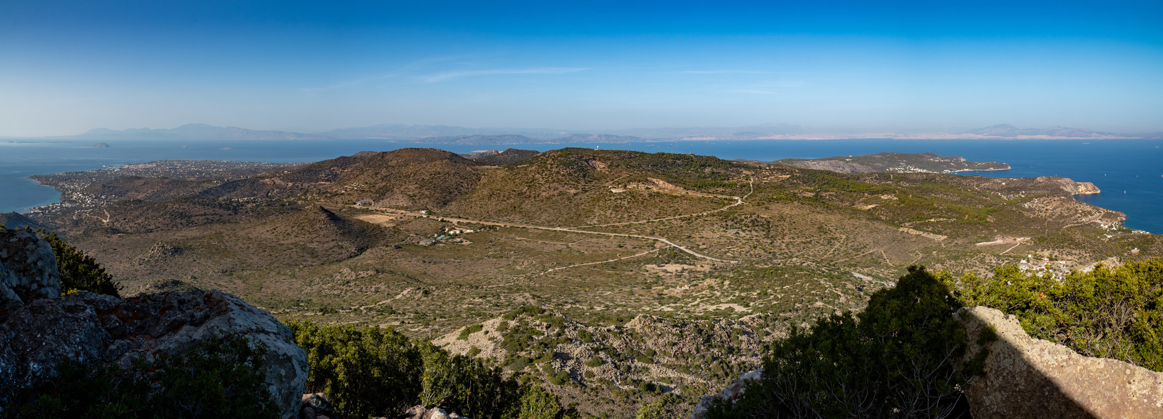 
Panoramic view from Mount Hellanion Oros in the north of the island of Aegina in the Saronic Gulf. To the left in the northwest is the town of Aegina. In the middle you can see a road leading from the west, from Pacheia Rachi across the island to the east. From this road there is a road to Lazarides to the north and a dirt road below Mount Oros to the south. There is a small parking lot at the intersection with the dirt road. In the background on the right you can see Athens on a clear day.