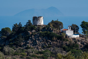 View of the old mill "Maltezou" and the church of St George from the car park below the mountain Hellanion Oros on the island of Aegina. In the background you can see the uninhabited island of Agios Georgios with a wind farm about 45 kilometres away.