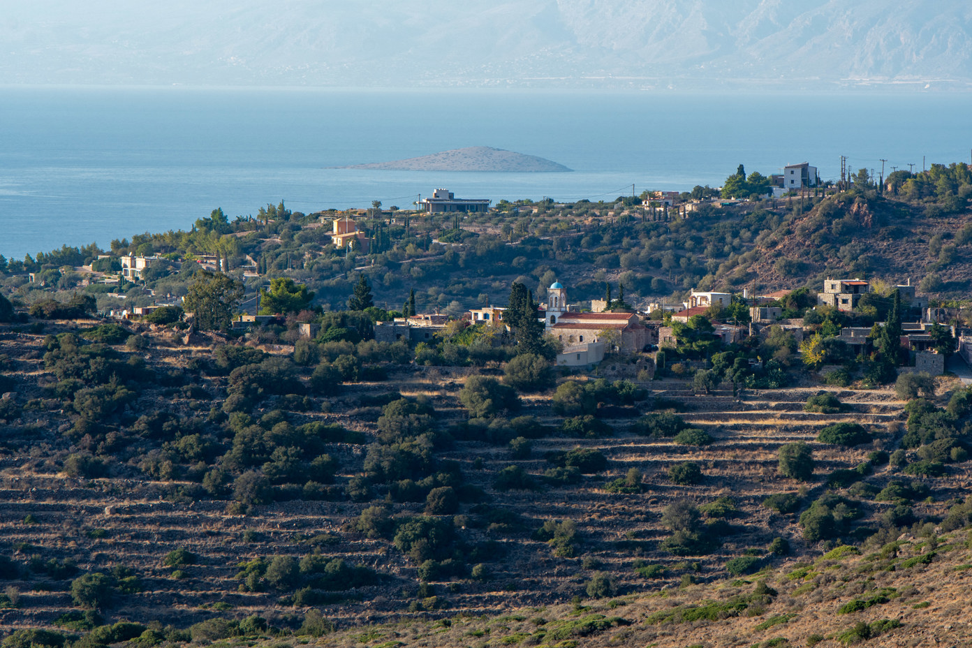 View from the hiking trail on Mount Ellanion Oros to Pachia Rachi with the church of Agios Dionysios above terraced fields on the island of Aegina. In the background, the Saronic Gulf with the small uninhabited island of Plateia (Πλατεία).