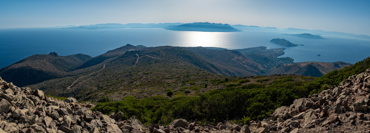 Panoramic view southwards over the Saronic Gulf from Mount Hellanion Oros on the island of Aegina. 
The south-east of the island is barely populated. In the middle you can see the end of the dirt road that leads from the car park north of the mountain. To the west is the town of Perdika on a headland and behind it the islands of Moni and Agkistri. In the background in the centre you can see the peninsula of the Methana volcano.