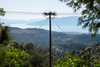 View from the country road near Pachia Rachi through the trees towards Perdika. In the foreground, a high-voltage power line parallel to the road. In the background a part of the Eleonas Valley, a high mountain valley with old olive trees.